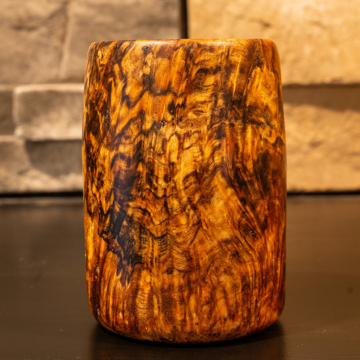 Shot glass made from Amboyna Burl wood against a stone fireplace background.