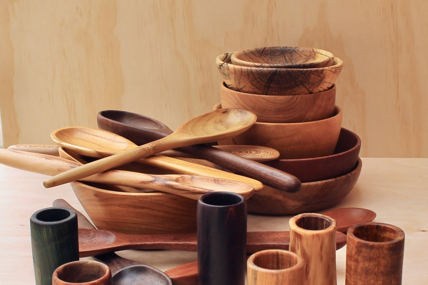 sets of hand crafted wooden shot glasses, espresso cups, bowls, spoons, and spatula on a light wooden background