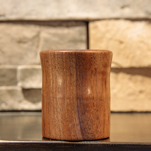 The Quiet Return Black Walnut shot glass, showing warm brown wood, contrasting vertical grain, a gently curved body, and a flared lip.