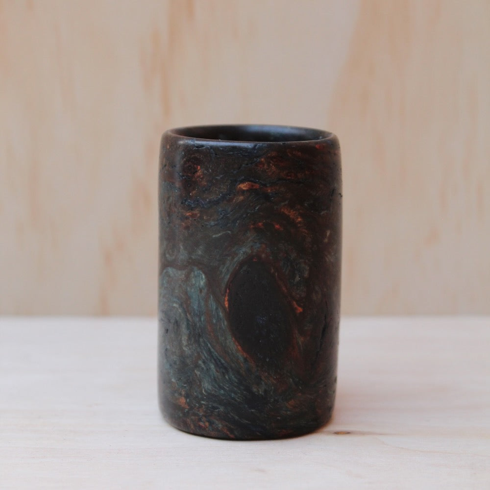 multi-color wooden shot glass made of exotic burl wood on a light wooden background. Rich dark browns flow into lighter golden streaks, with irregular swirls and knots that create depth across the cylindrical walls