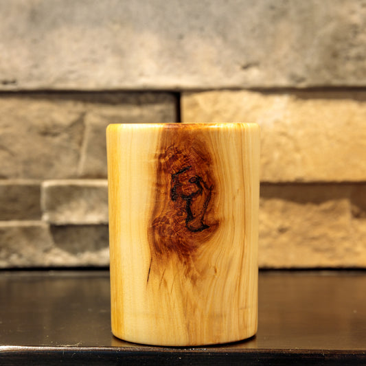 Cylindrical Narrowleaf Cottonwood wooden shot glass with pale golden grain and a prominent reddish-brown knot with dark, irregular markings on its front, photographed against a stone background.