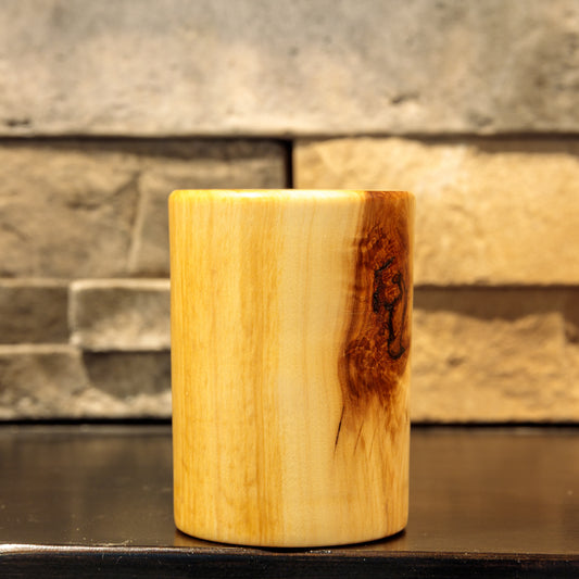 Side rotation of a cylindrical Narrowleaf Cottonwood shot glass, showing the dark reddish-brown knot near the right edge and long, flowing golden grain across the pale wood.