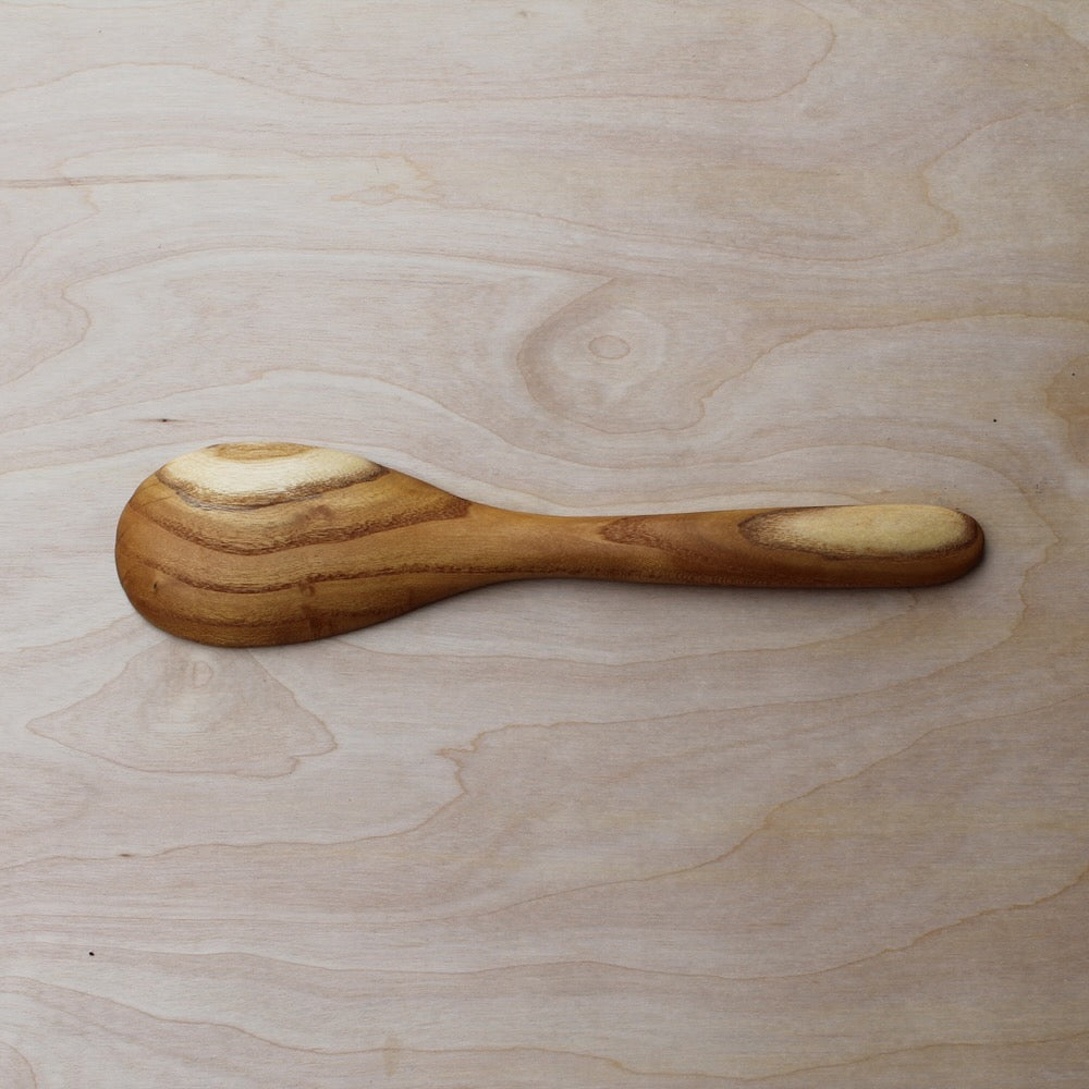 back of a hand made wooden spatula with dark brown stripes on a light wooden surface. pattern consists of butter yellow wood mixed with light brown wood