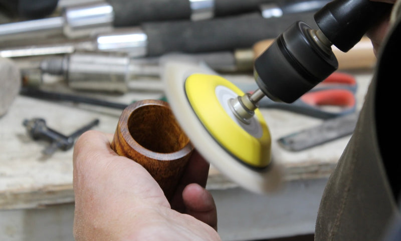 Hand holding a wooden object near a polishing tool with a yellow pad on a workshop background.